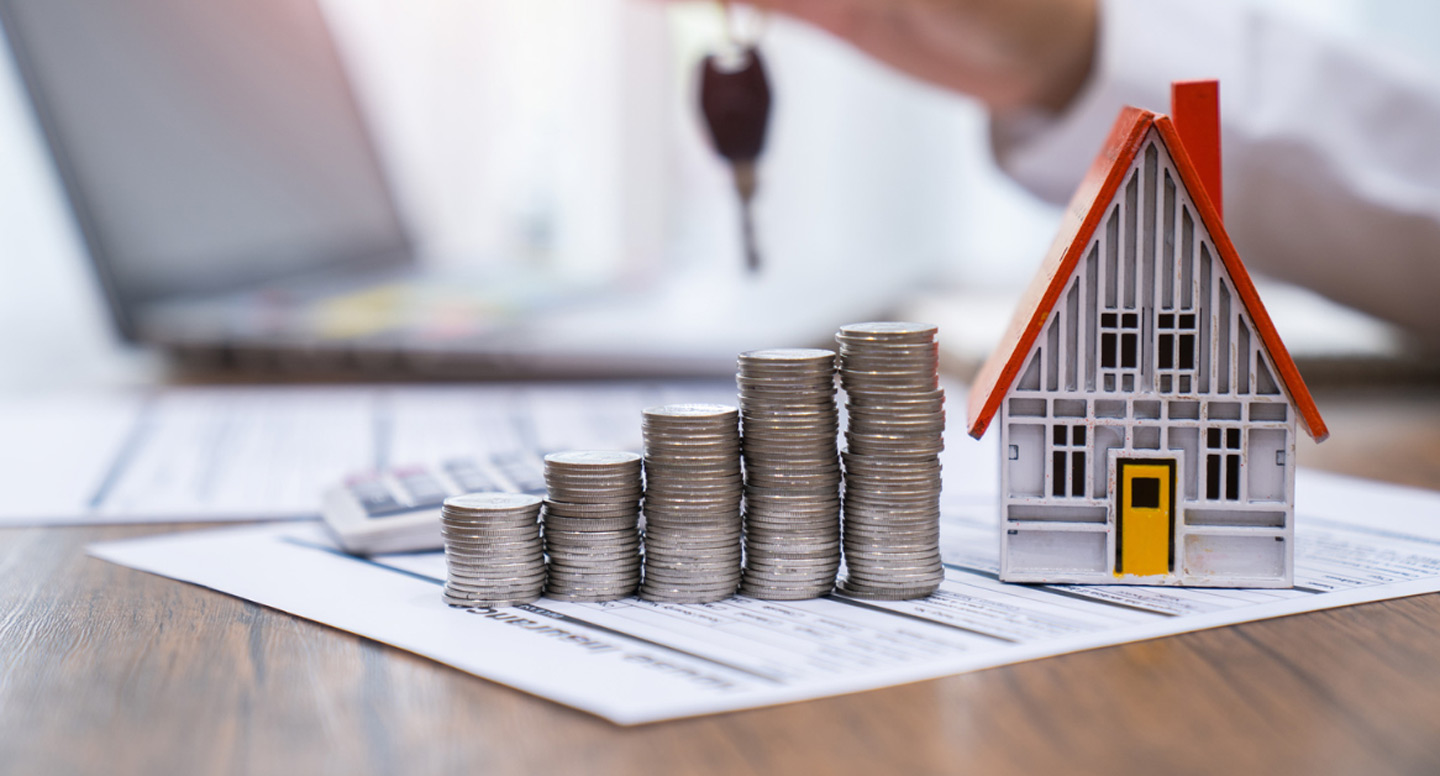 A model house with a red roof sits beside stacked coins on financial documents, symbolizing real estate investment. A hand holds car keys in the background.