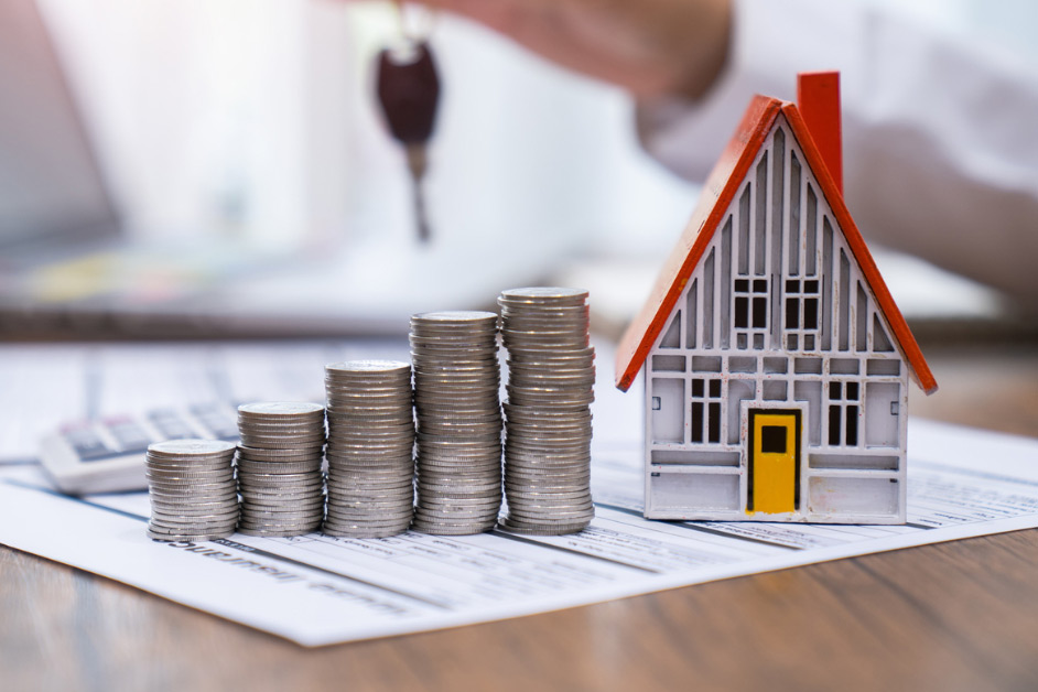 A model house with a red roof sits beside stacked coins on financial documents, symbolizing real estate investment. A hand holds car keys in the background.
