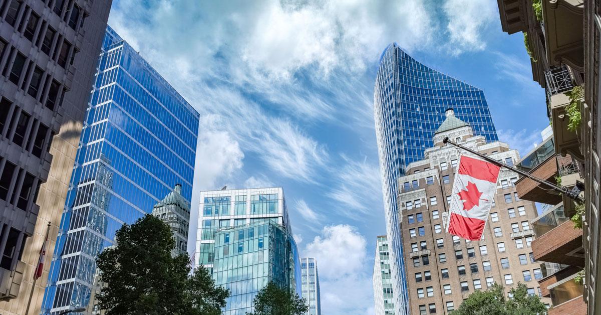 Downtown Toronto skyline with modern glass skyscrapers, historic buildings, and a large Canadian flag beneath a bright, cloud-streaked sky.