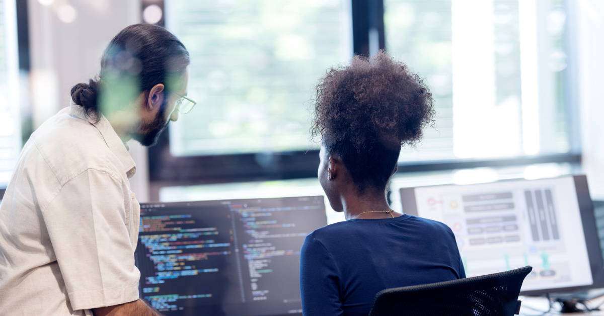 A bearded developer and a colleague review colorful code on dual monitors in a bright office, sharing a focused, collaborative moment.