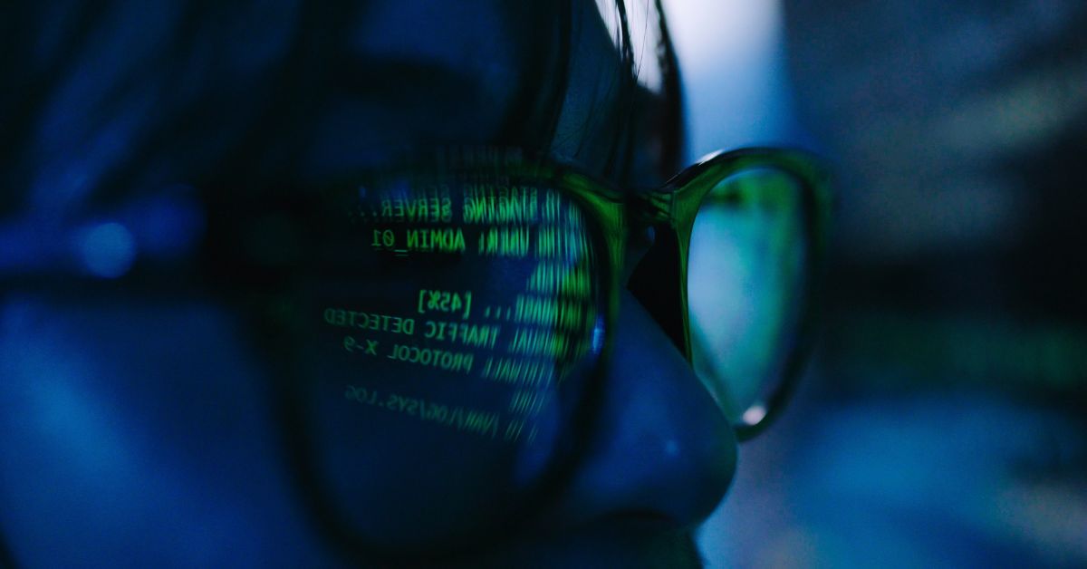 Close-up of a person wearing glasses, green computer code reflected in one lens against a dark blue background, creating a moody cyber-tech feel.