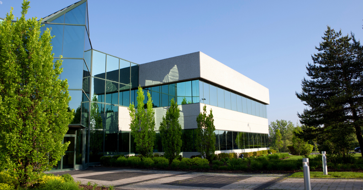 Office building with reflective glass facade, surrounded by lush green trees and shrubs under a clear blue sky