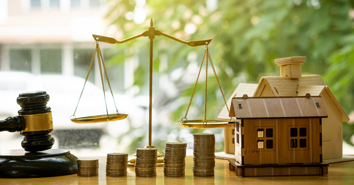 A judge's gavel, balance scale, stacked coins, and a wooden house model sit on a table, symbolizing real estate finance and legal decision-making.
