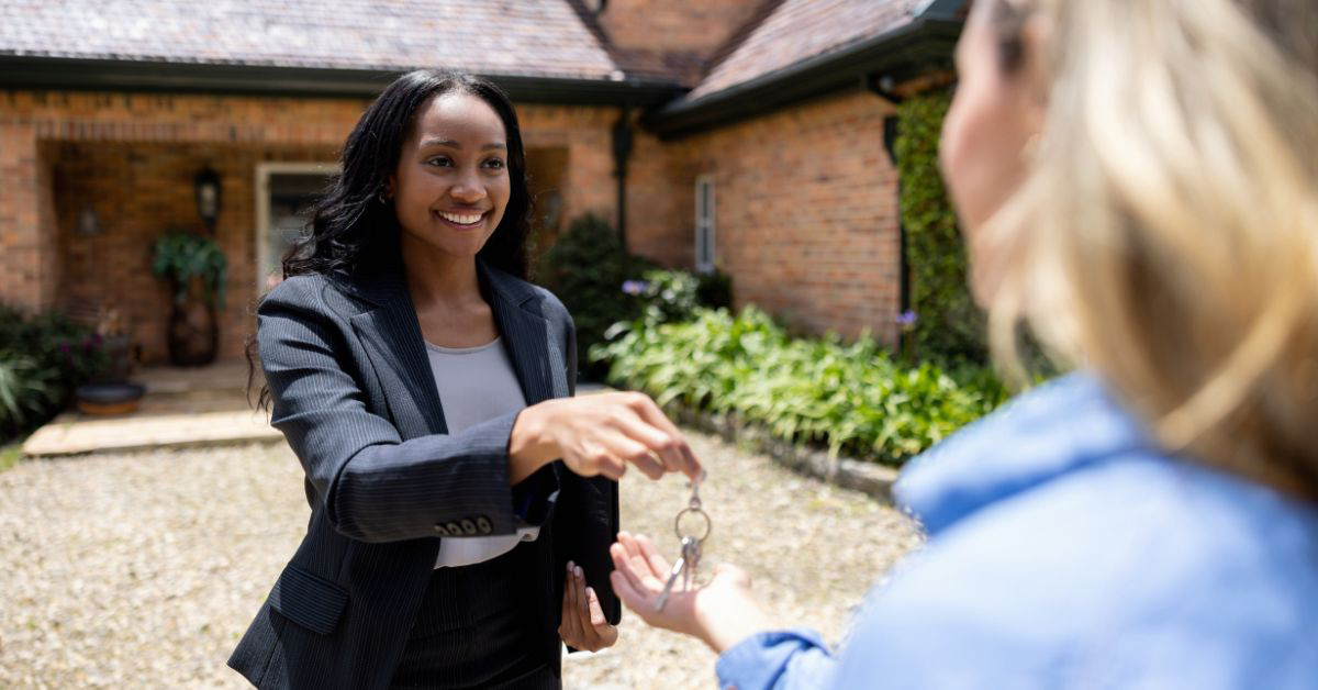 A female Real Estate Agent hands house keys to another person outside a brick home.