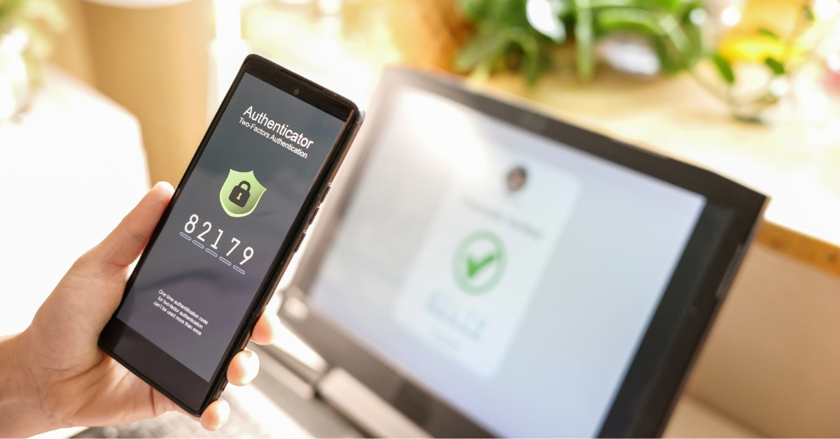 A hand holds a smartphone displaying an authenticator app and six-digit security code beside a blurred laptop login screen, suggesting secure online access.