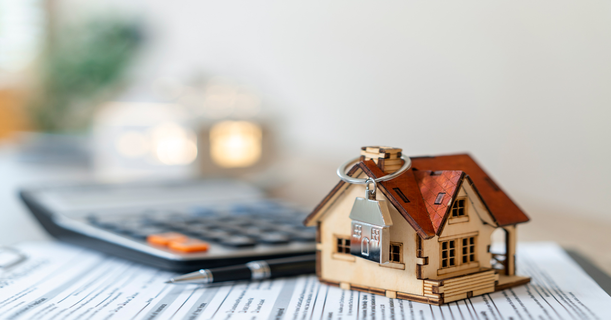 A small wooden house model with a key sits on loan documents next to a pen and calculator.