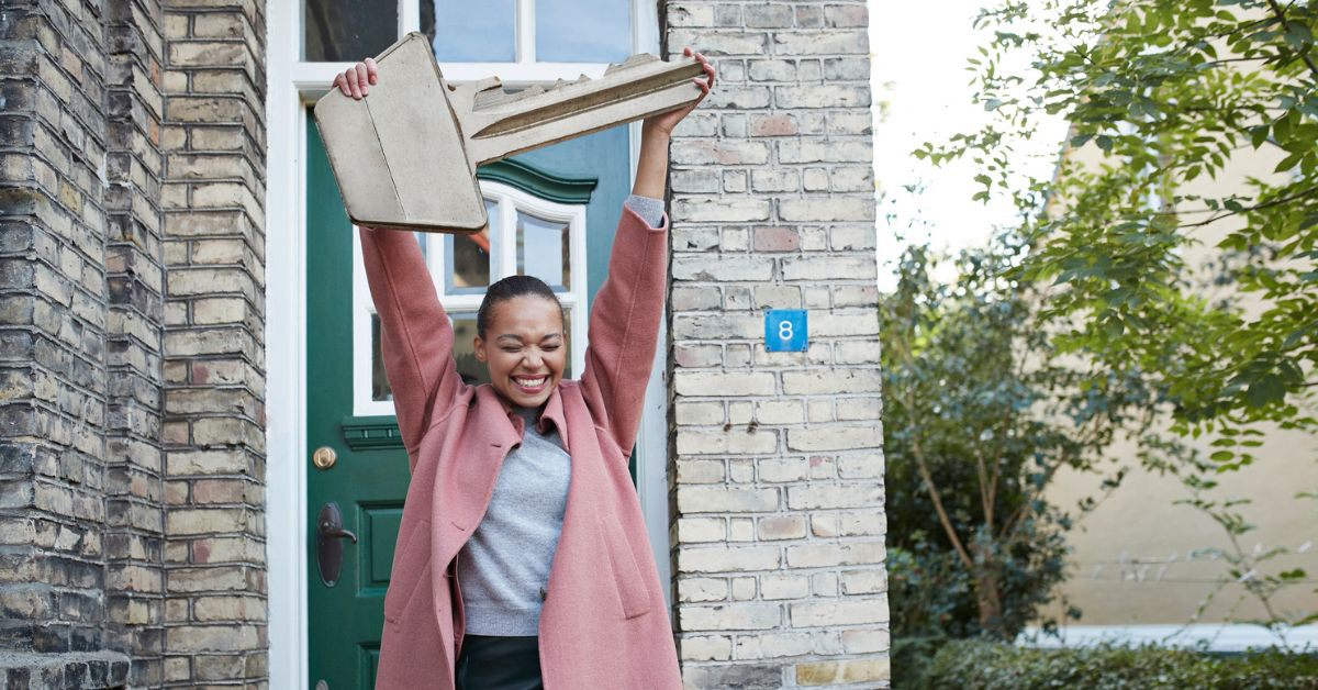 A joyful woman in a pink coat holds a large cardboard key above her head, conveying excitement and success after buying new home.