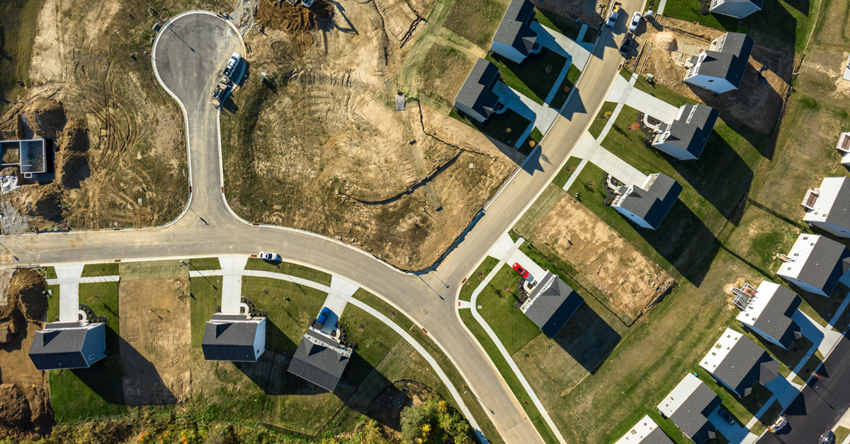 Aerial view of a suburban housing development with curving roads, new homes, vacant lots, and construction equipment in daylight.