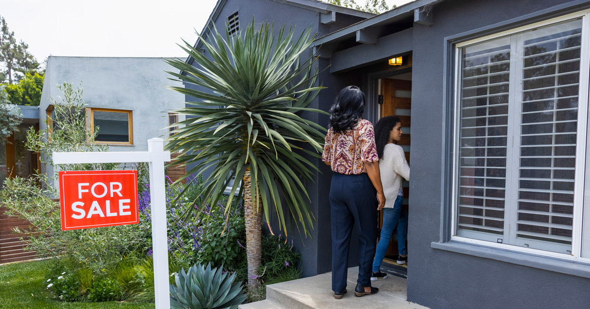 A red “FOR SALE” sign stands outside a modern gray home as two women enter, framed by lush yucca plants and a bright, welcoming doorway.