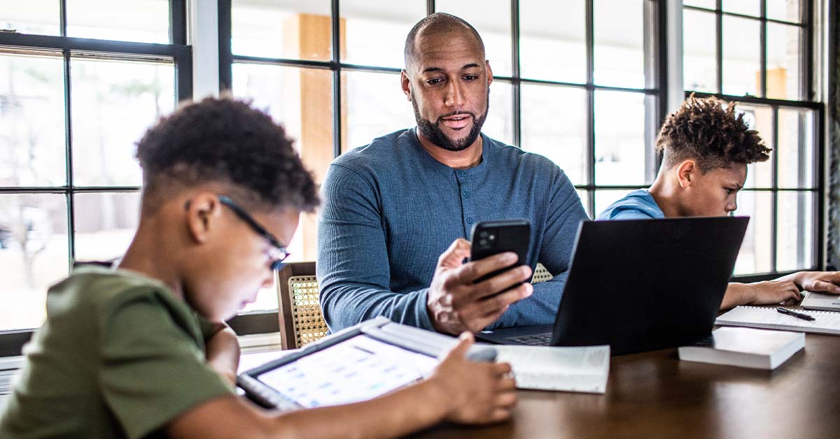 Father working from home in residential kitchen with family in background, all using electronic devices.
