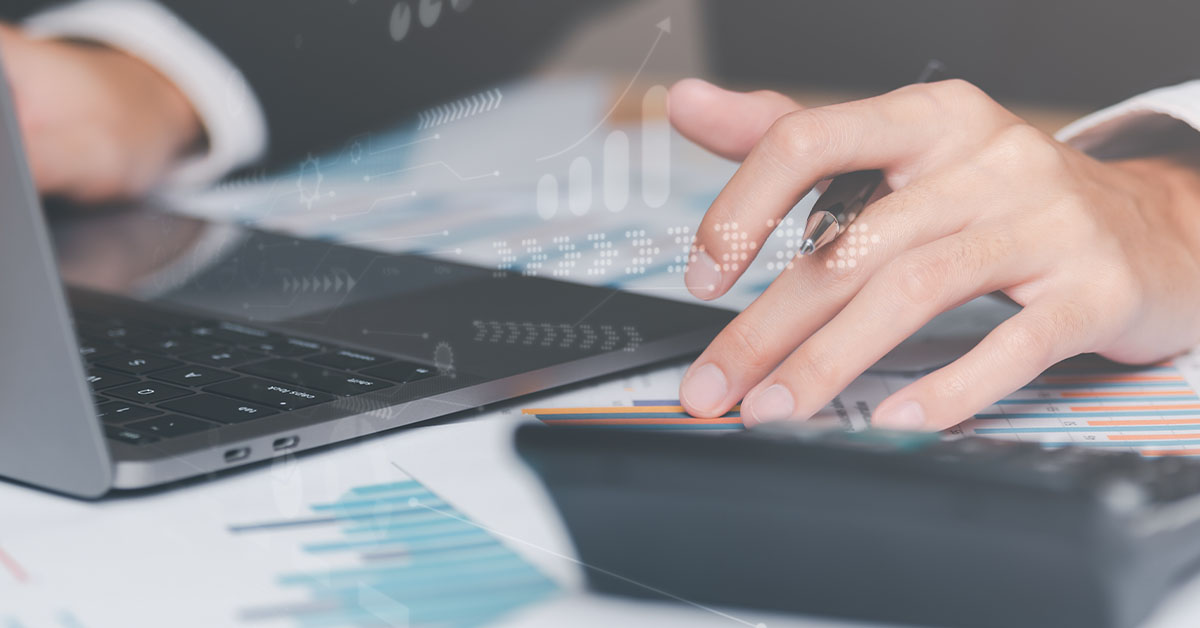 Image A young business man, using a laptop and calculator, on a busy desk covered with graphs and charts.